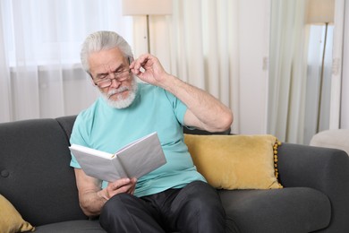 Photo of Senior man with vision problem reading book on sofa at home