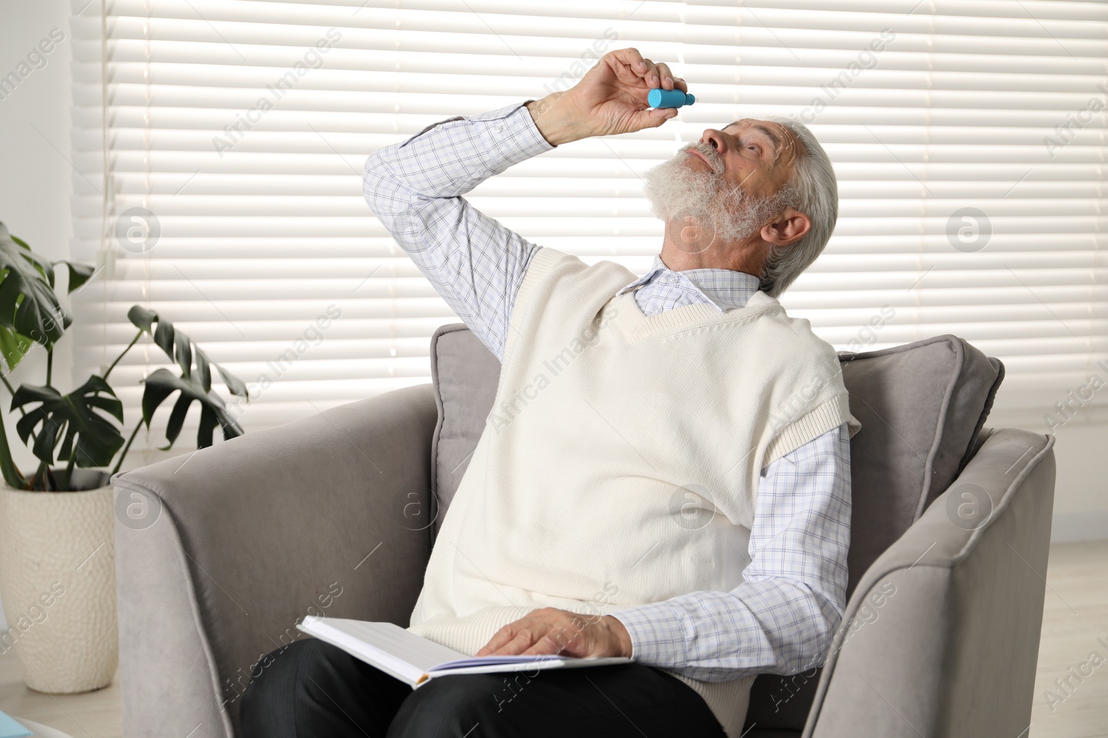 Senior man applying eye drops in armchair at home Photo of Senior man applying eye drops in armchair at home