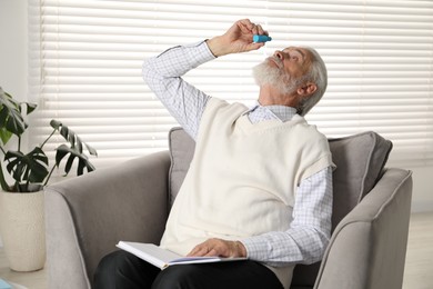 Photo of Senior man applying eye drops in armchair at home