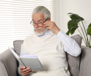 Photo of Senior man with vision problem reading book in armchair at home