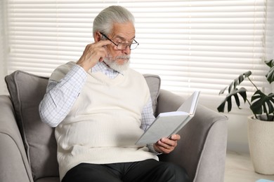 Photo of Senior man with vision problem reading book in armchair at home