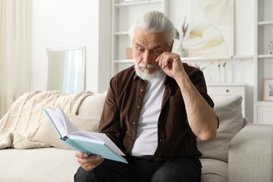 Photo of Senior man with vision problem reading book on sofa at home