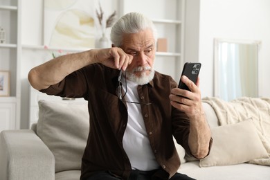 Photo of Senior man with vision problem using smartphone on sofa at home