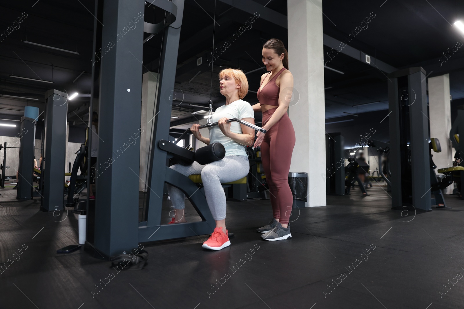 Senior woman training on lat pulldown machine supervised by professional trainer in gym Photo of Senior woman training on lat pulldown machine supervised by professional trainer in gym