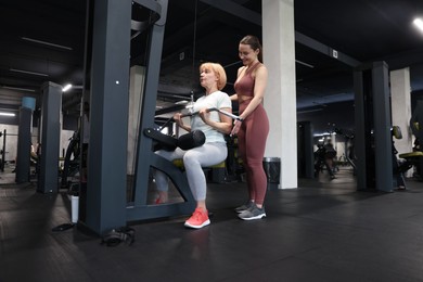 Photo of Senior woman training on lat pulldown machine supervised by professional trainer in gym