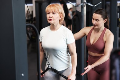 Photo of Senior woman training on pulley machine supervised by professional trainer in gym