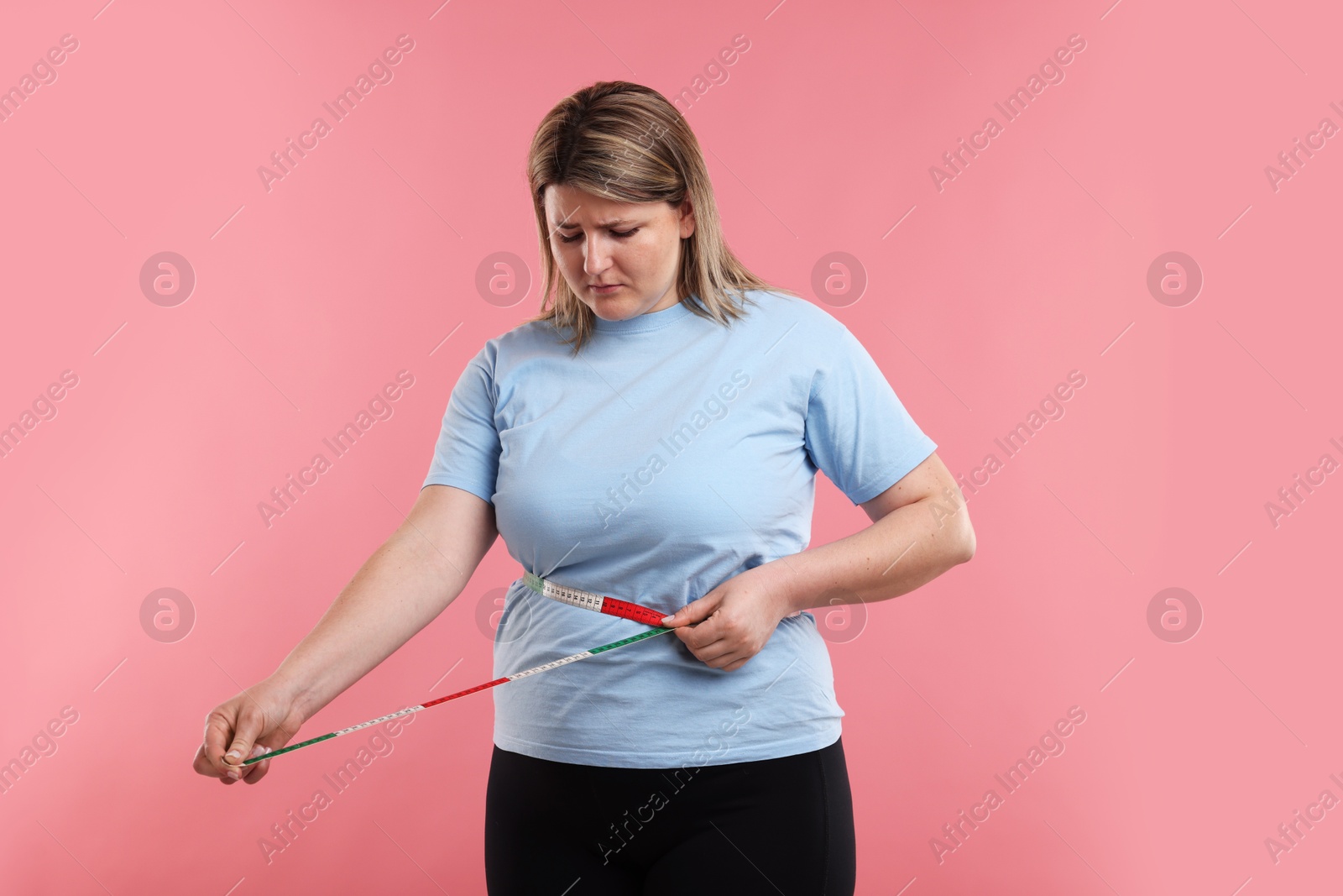 Weight loss. Plus size woman measuring waist with tape on pink background Photo of Weight loss. Plus size woman measuring waist with tape on pink background