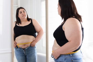 Photo of Overweight woman measuring her belly with tape near mirror indoors