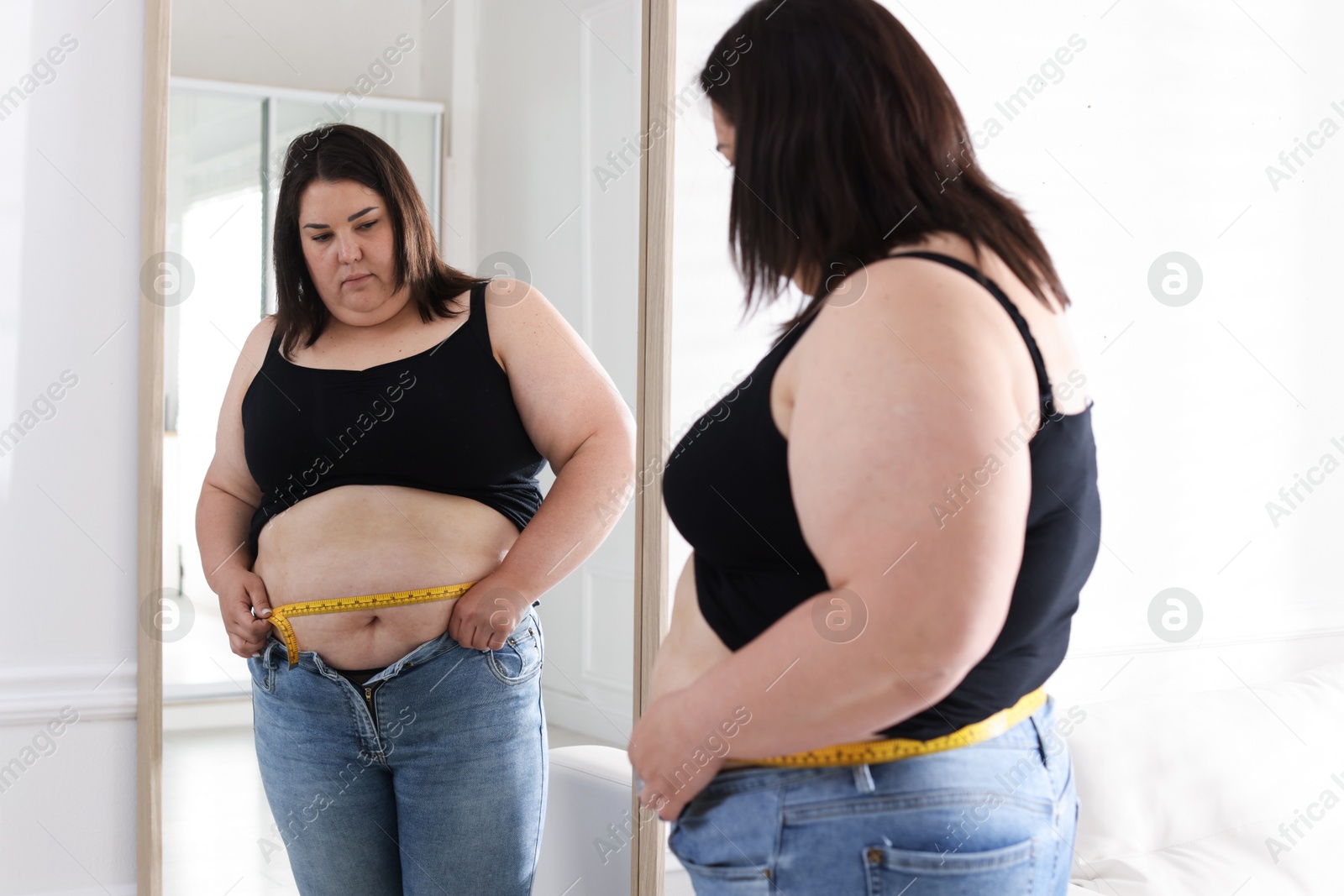 Overweight woman measuring her belly with tape near mirror indoors Photo of Overweight woman measuring her belly with tape near mirror indoors