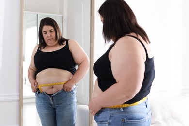 Photo of Overweight woman measuring her belly with tape near mirror indoors