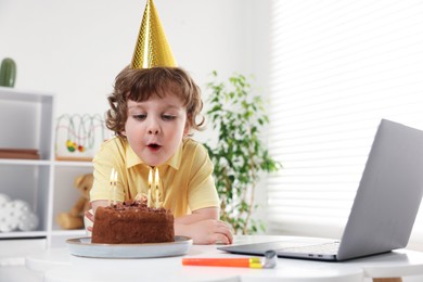 Photo of Birthday celebration. Little boy blowing candles on cake while having video call via laptop at table indoors. Space for text