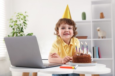 Photo of Birthday celebration. Little boy with cake having video call via laptop at table indoors