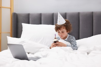 Photo of Birthday celebration. Little boy with cupcake having video call via laptop in bed at home