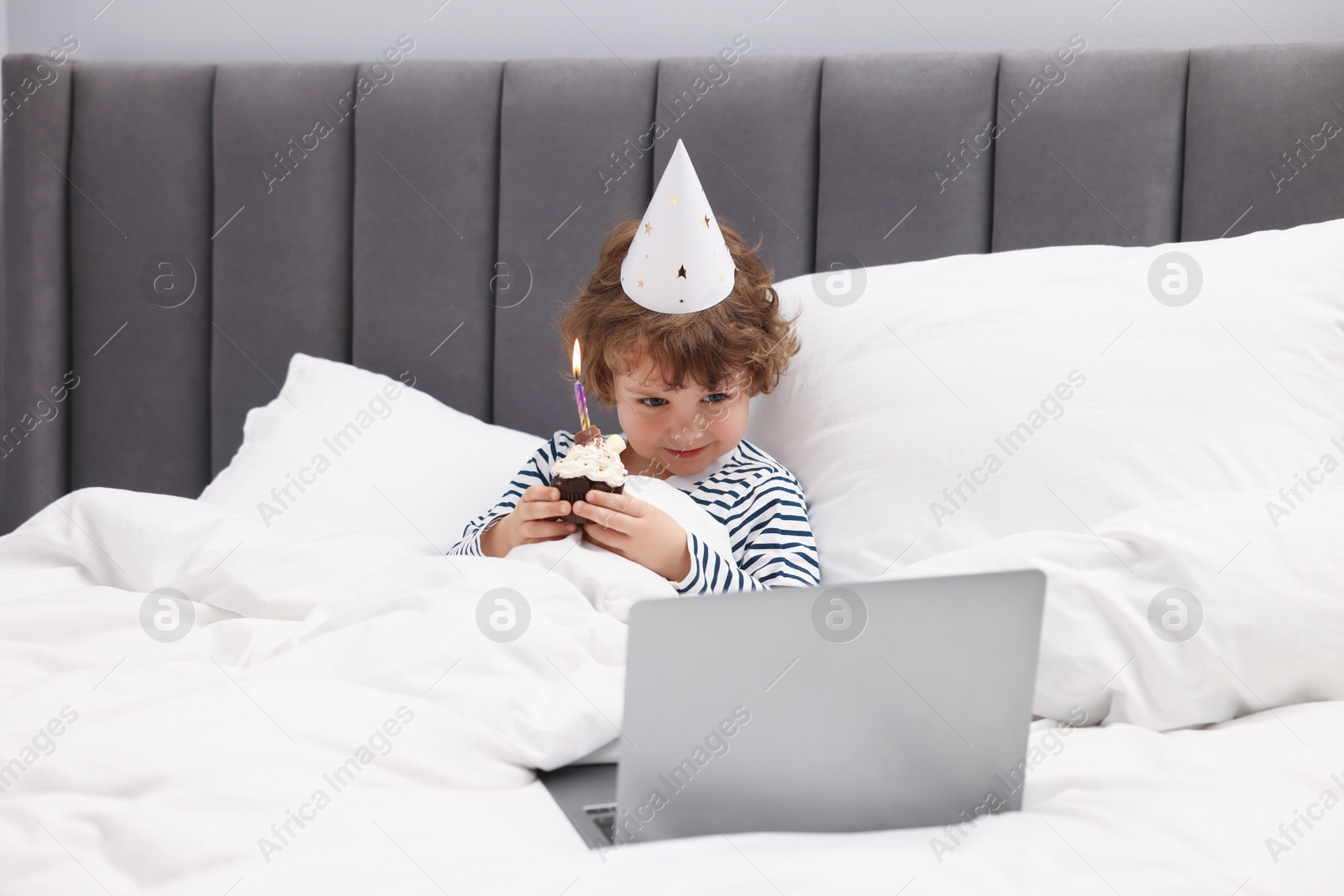Birthday celebration. Little boy with cupcake having video call via laptop in bed at home Photo of Birthday celebration. Little boy with cupcake having video call via laptop in bed at home