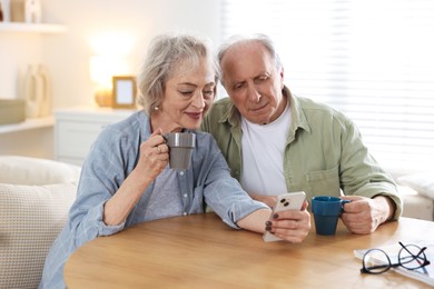 Photo of Lovely senior couple enjoying hot drinks while watching something on smartphone at table indoors