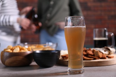 Photo of Glass of beer with snacks on table and friends chatting indoors, selective focus