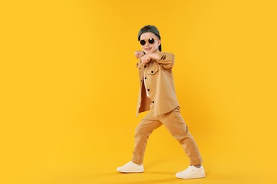 Photo of Happy little boy in sunglasses and baseball hat dancing on orange background. Space for text