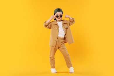 Photo of Happy little boy in sunglasses and baseball hat dancing on orange background