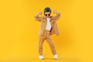 Photo of Happy little boy in sunglasses and baseball hat dancing on orange background