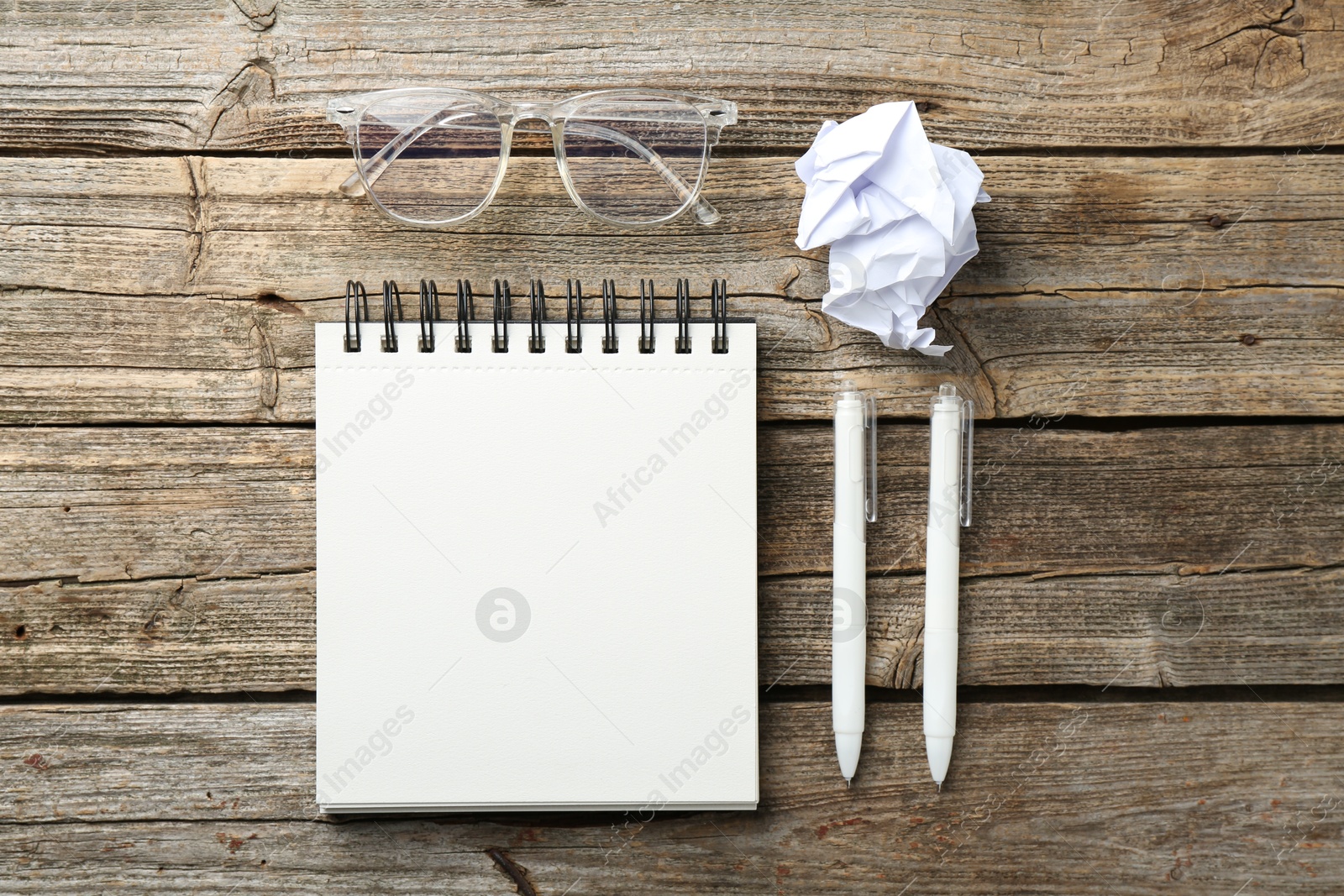 Photo of Open blank notebook, glasses, crumpled paper and pens on wooden table, flat lay