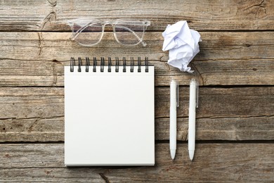 Photo of Open blank notebook, glasses, crumpled paper and pens on wooden table, flat lay