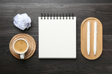 Photo of Open blank notebook, coffee, crumpled paper and pens on black wooden table, flat lay