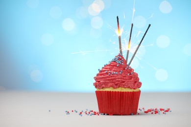 Photo of Burning sparklers in cupcake and sprinkles on white table against light blue background with blurred lights, space for text
