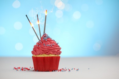Photo of Burning sparklers in cupcake and sprinkles on white table against light blue background with blurred lights, space for text