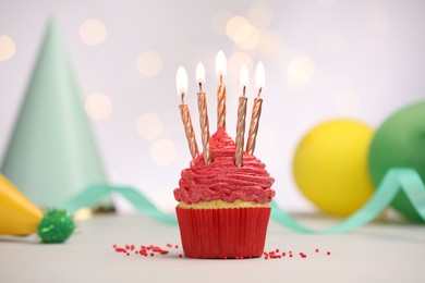 Photo of Burning candles in cupcake and sprinkles on white table against blurred lights
