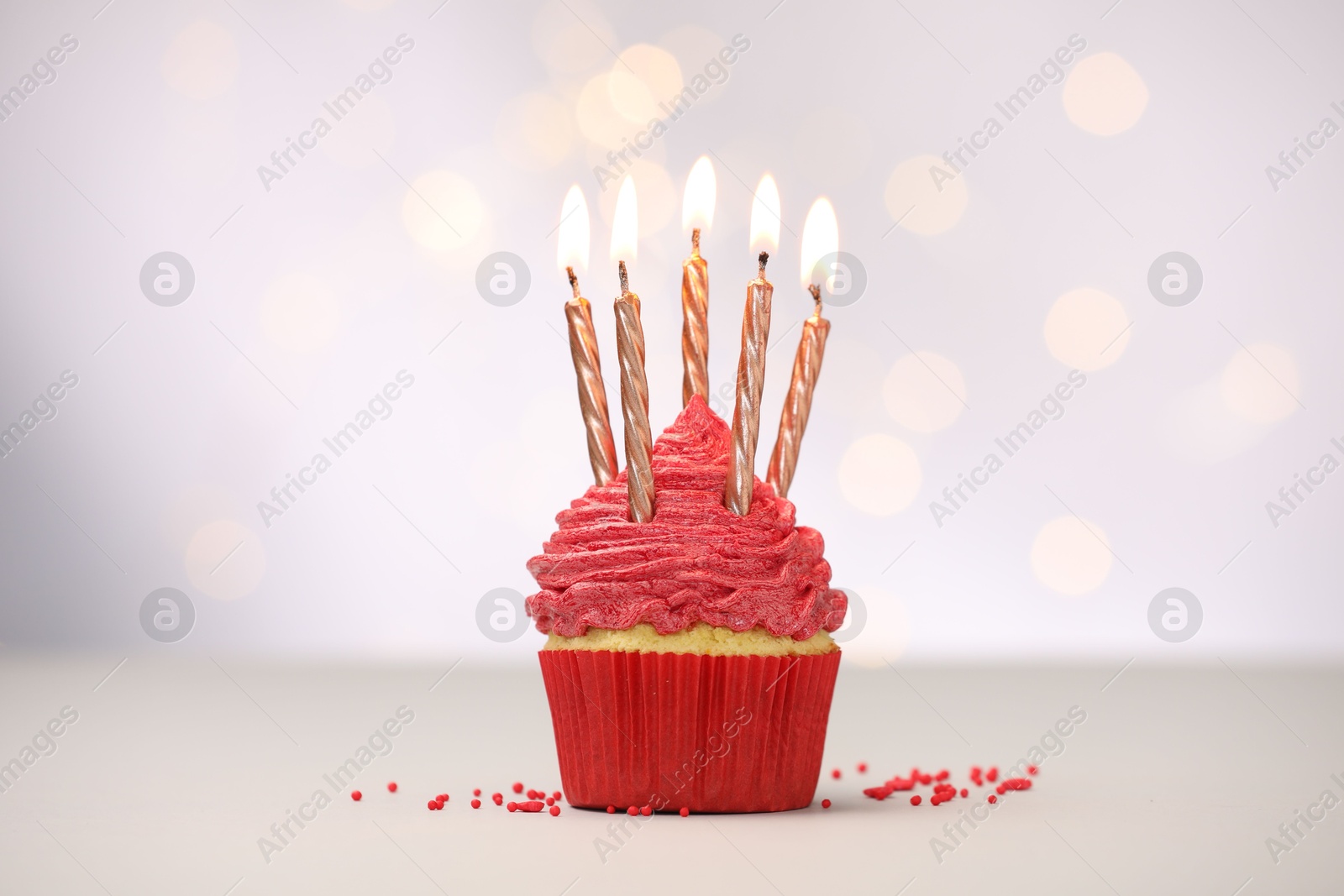 Photo of Burning candles in cupcake and sprinkles on white table against blurred lights