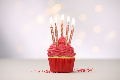 Photo of Burning candles in cupcake and sprinkles on white table against blurred lights