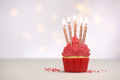 Photo of Burning candles in cupcake and sprinkles on white table against blurred lights, space for text