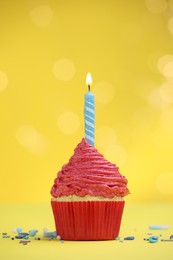 Photo of Burning candle in cupcake and sprinkles on yellow background with blurred lights
