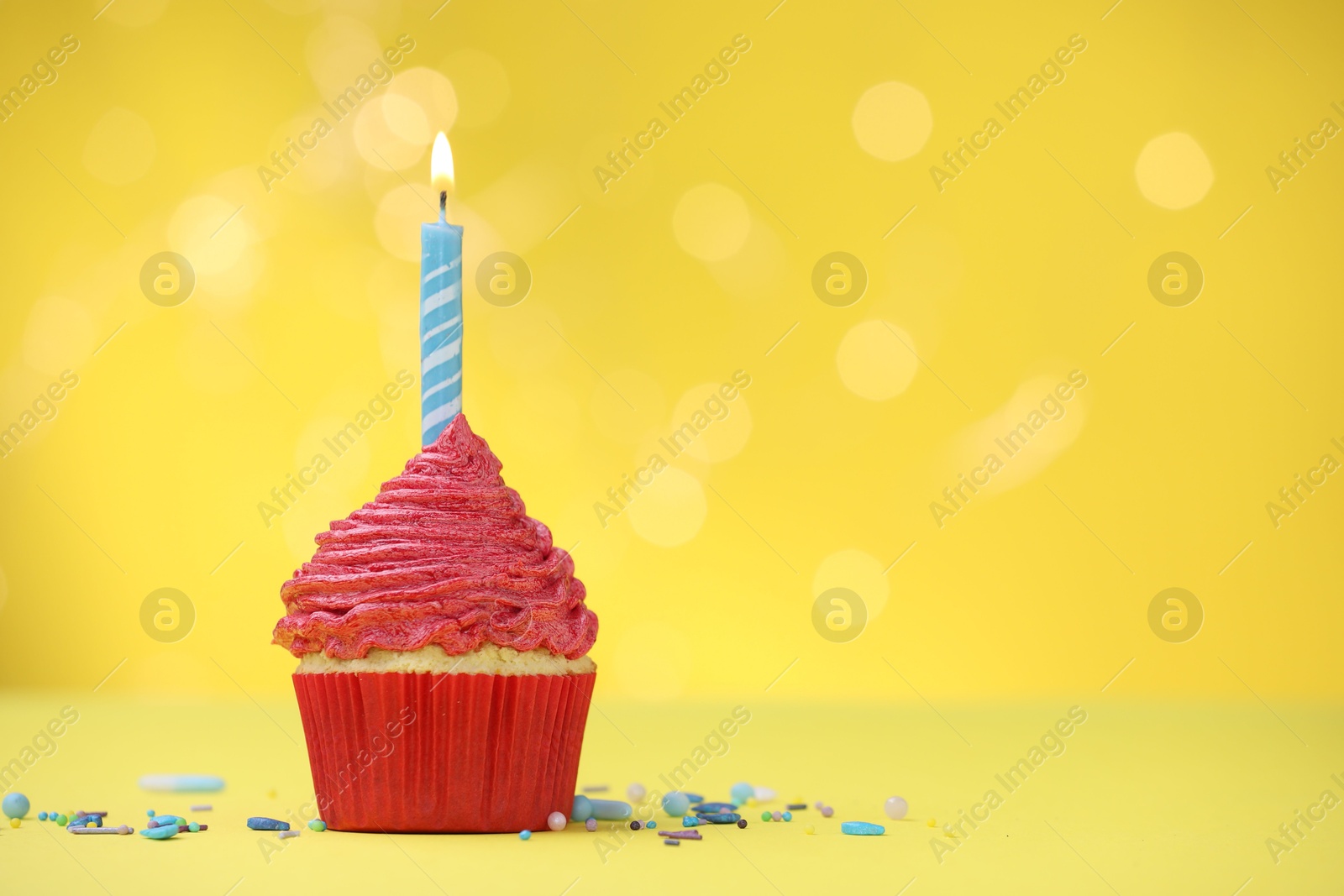 Burning candle in cupcake and sprinkles on yellow background with blurred lights, space for text Photo of Burning candle in cupcake and sprinkles on yellow background with blurred lights, space for text