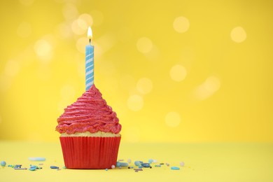 Photo of Burning candle in cupcake and sprinkles on yellow background with blurred lights, space for text