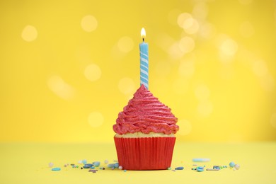 Photo of Burning candle in cupcake and sprinkles on yellow background with blurred lights