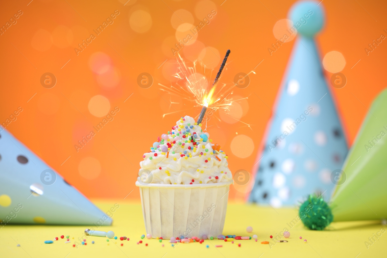Burning sparkler in cupcake, sprinkles and party decor on yellow table against orange background blurred lights Photo of Burning sparkler in cupcake, sprinkles and party decor on yellow table against orange background blurred lights