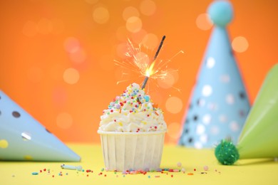 Photo of Burning sparkler in cupcake, sprinkles and party decor on yellow table against orange background blurred lights