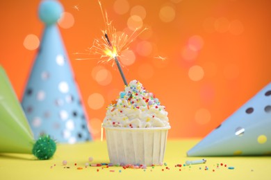 Photo of Burning sparkler in cupcake, sprinkles and party decor on yellow table against orange background blurred lights
