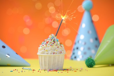Photo of Burning sparkler in cupcake, sprinkles and party decor on yellow table against orange background blurred lights