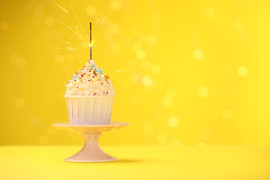 Photo of Burning sparkler in cupcake, sprinkles and stand on yellow background with blurred lights, space for text