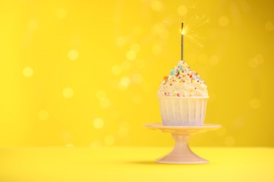 Photo of Burning sparkler in cupcake, sprinkles and stand on yellow background with blurred lights, space for text