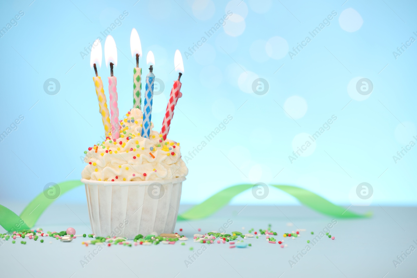 Photo of Burning candles in cupcake and sprinkles on white table against light blue background with blurred lights