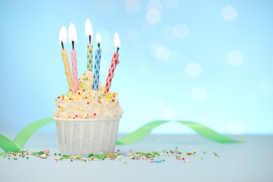 Photo of Burning candles in cupcake and sprinkles on white table against light blue background with blurred lights