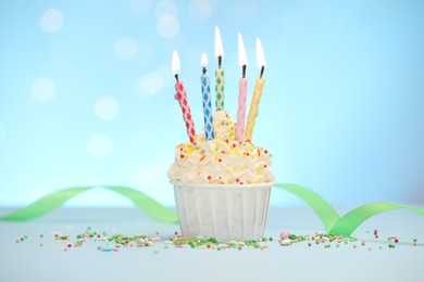 Photo of Burning candles in cupcake and sprinkles on white table against light blue background with blurred lights