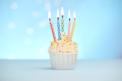 Photo of Burning candles in cupcake and sprinkles on white table against light blue background with blurred lights