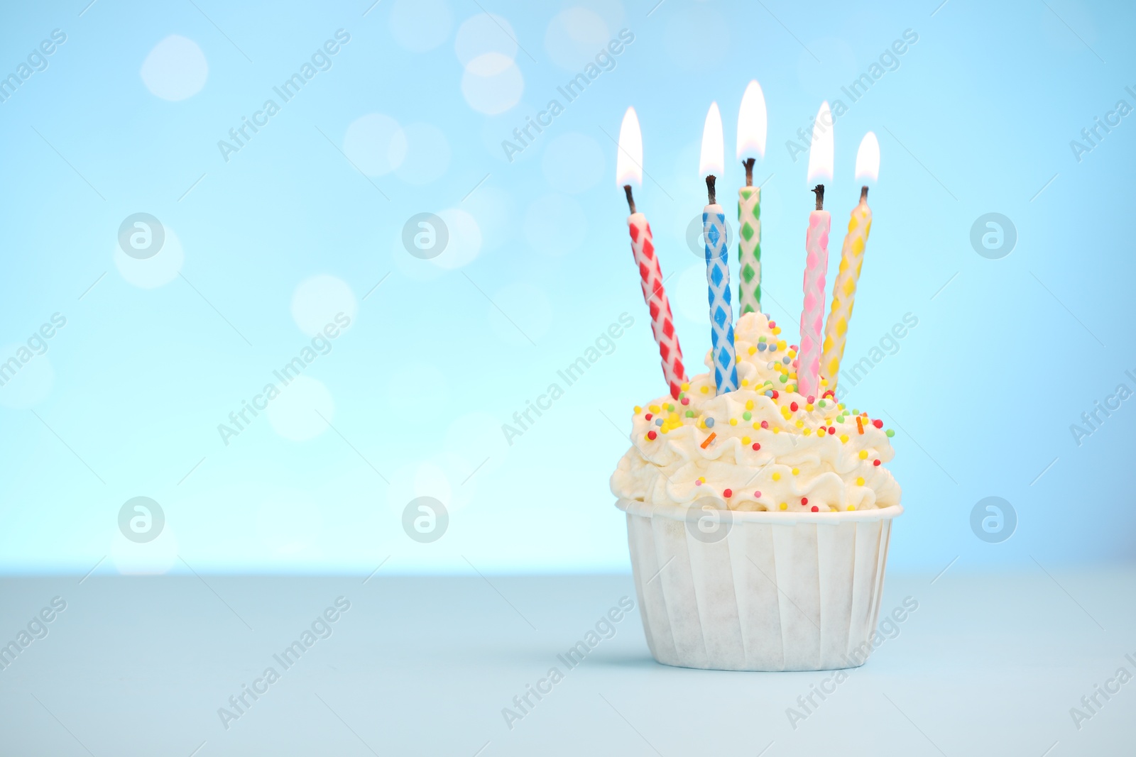 Photo of Burning candles in cupcake and sprinkles on white table against light blue background with blurred lights, space for text