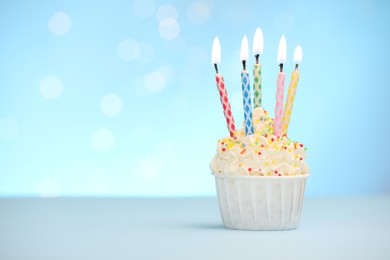 Photo of Burning candles in cupcake and sprinkles on white table against light blue background with blurred lights, space for text