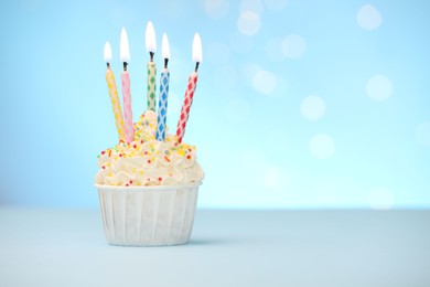 Photo of Burning candles in cupcake and sprinkles on white table against light blue background with blurred lights, space for text