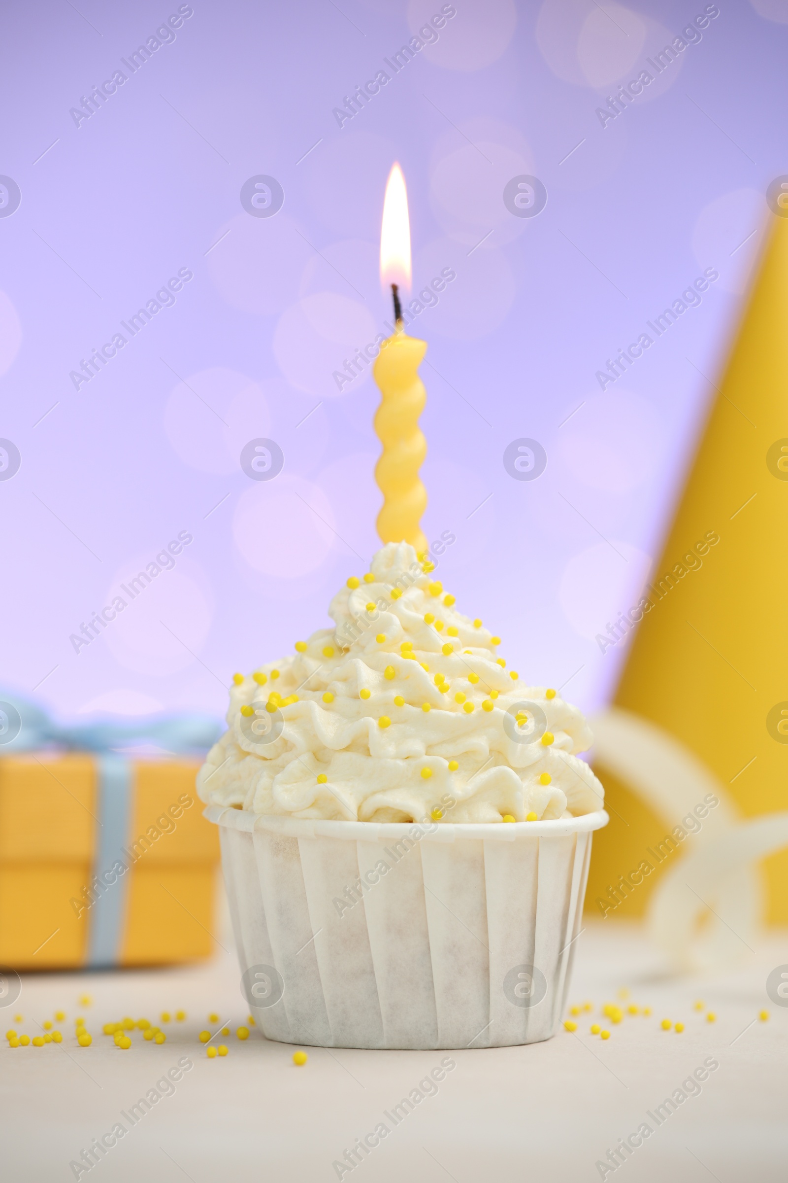 Photo of Burning candle in cupcake, sprinkles and party decor on white table against violet background with blurred lights